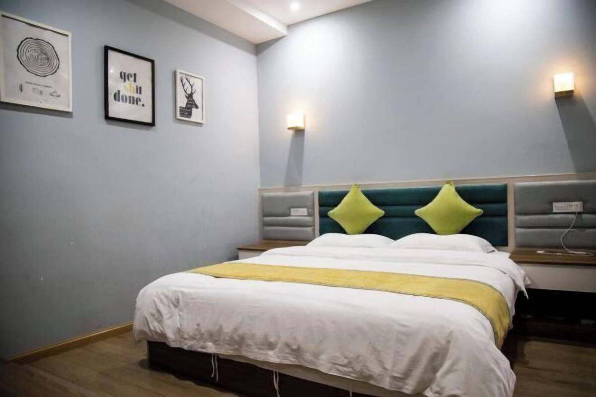 A neatly made bed with a white and yellow bedspread, a teal headboard, and two green pillows. There are framed pictures on the wall and a bedside lamp.