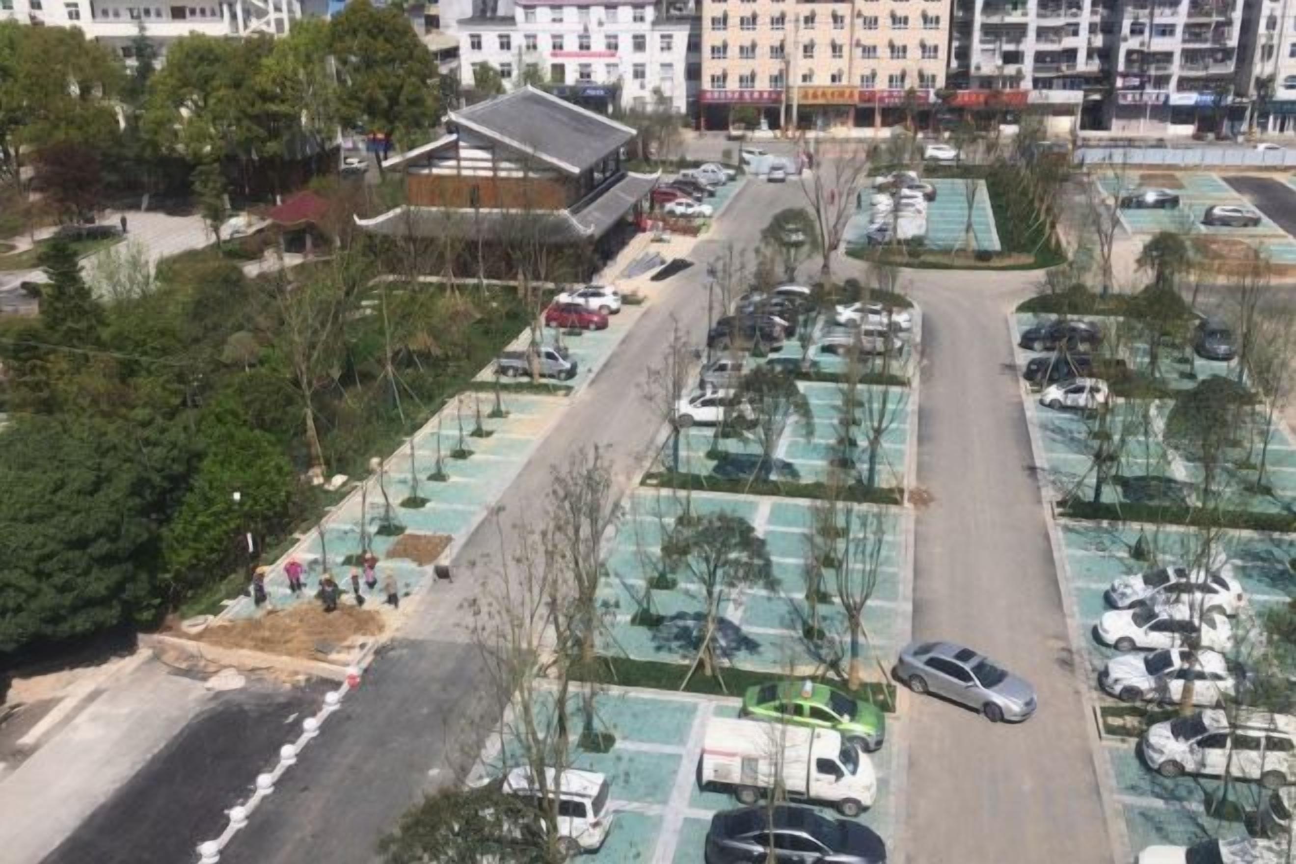 An aerial view of a parking lot with multiple rows of parked cars, a central pedestrian pathway, and surrounding greenery.