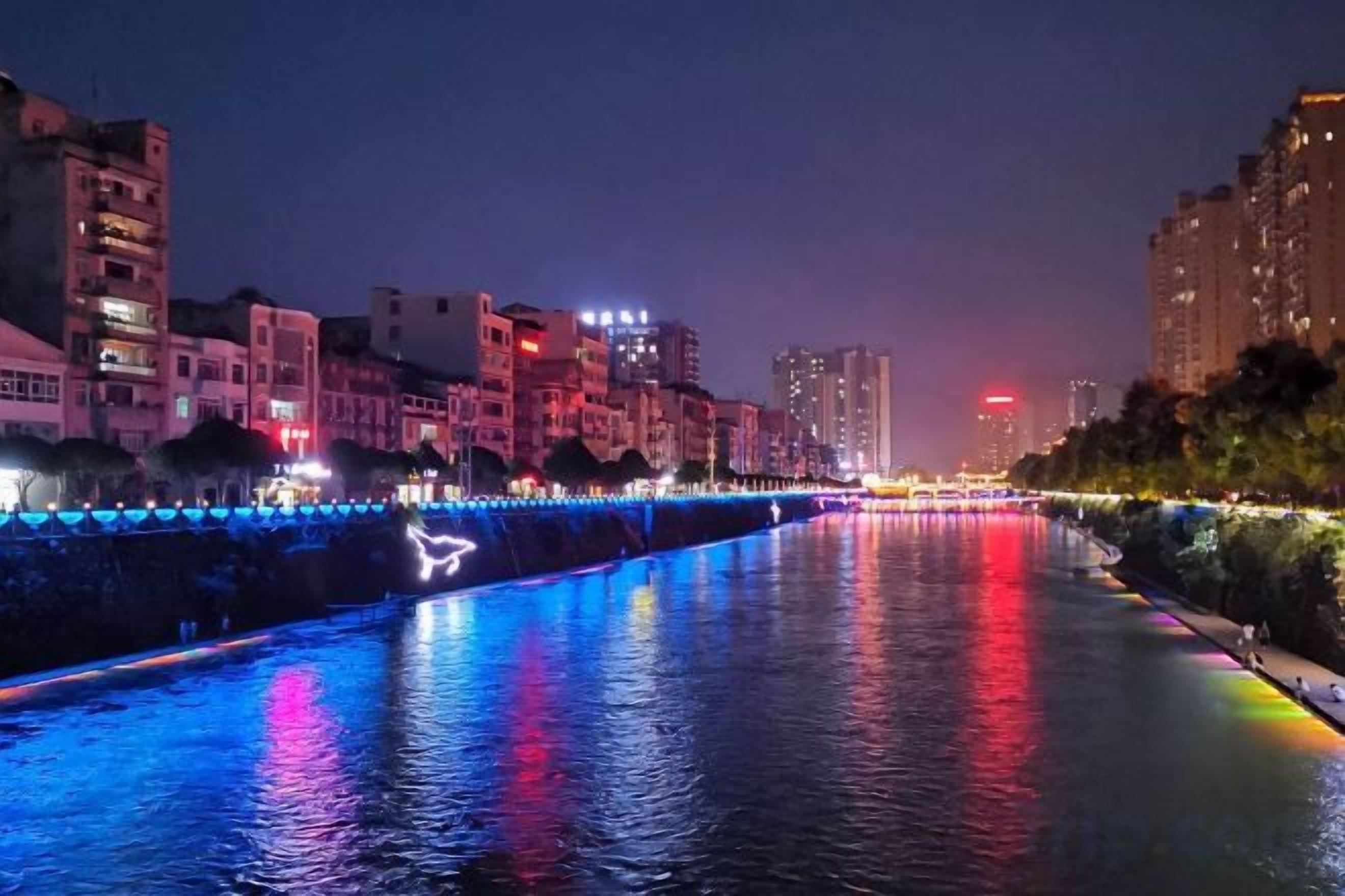 A nighttime cityscape with a river reflecting colorful lights, buildings along the riverbank, and a distant red light in the skyline.