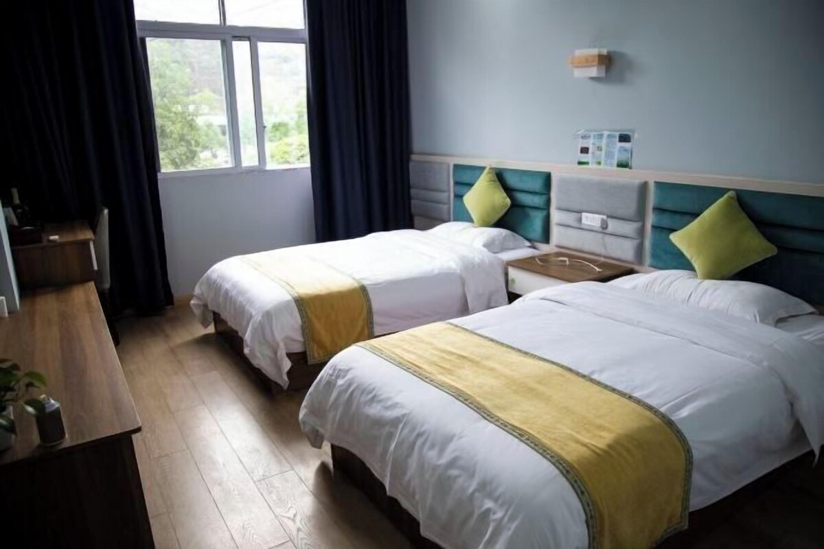 Une chambre d’hôtel avec deux lits, un bureau en bois, une fenêtre avec des rideaux et une petite table sur laquelle est posée une plante.