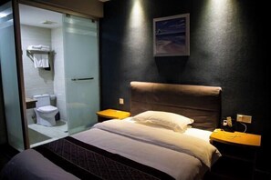 Une chambre d’hôtel moderne dotée d’une douche à parois de verre, d’un grand lit avec du linge de lit blanc et d’un mur foncé orné d’un tableau encadré.