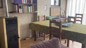 Ein Zimmer mit Esstisch, bedeckt mit einer grünen Tischdecke, Holzsesseln, einem violetten Sofa, einem Bücherregal mit Büchern und einem schwarzen Heizgerät.
