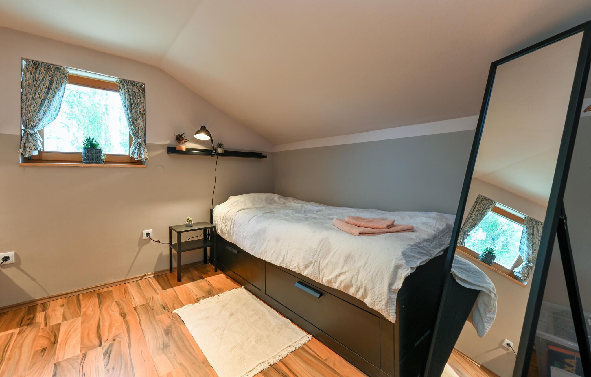 Un dormitorio con cama, mesita de noche, lámpara, ventana con cortinas y espejo.