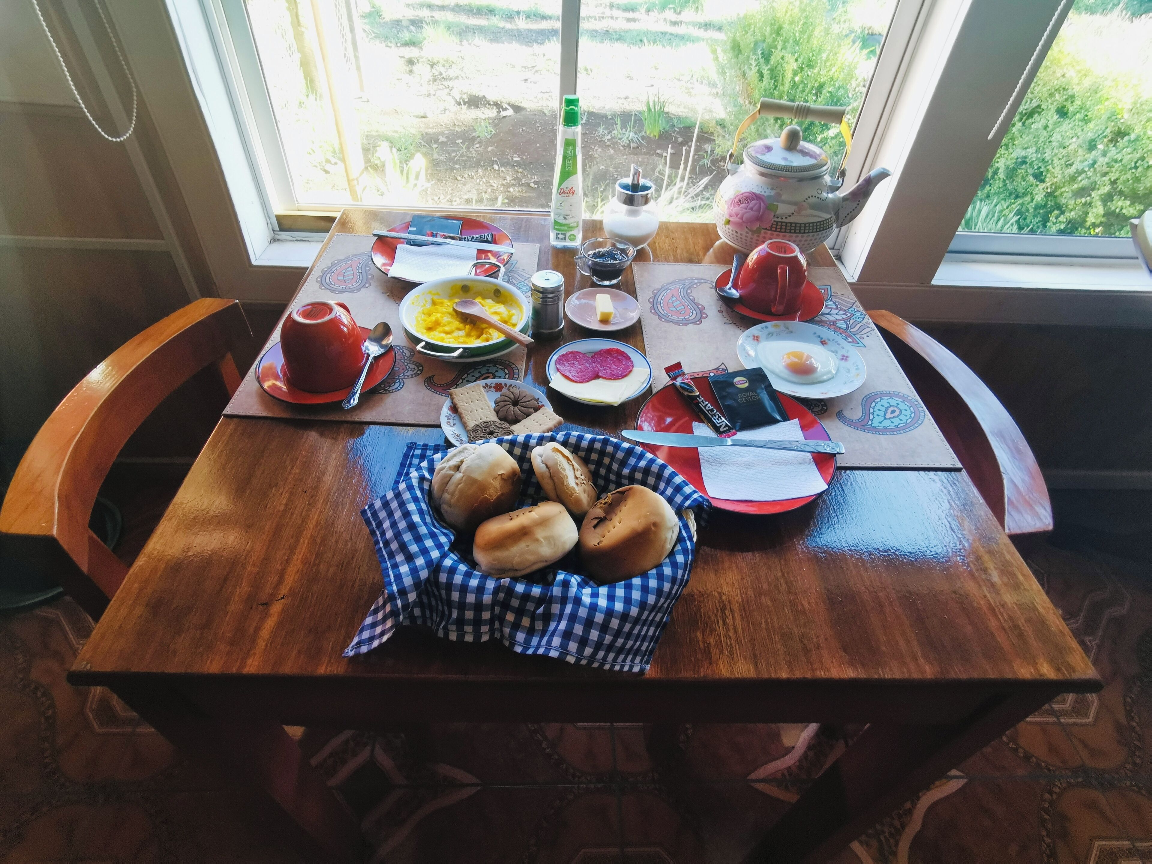 Een ontbijttafel met diverse gerechten, een theepot en uitzicht op een tuin.
