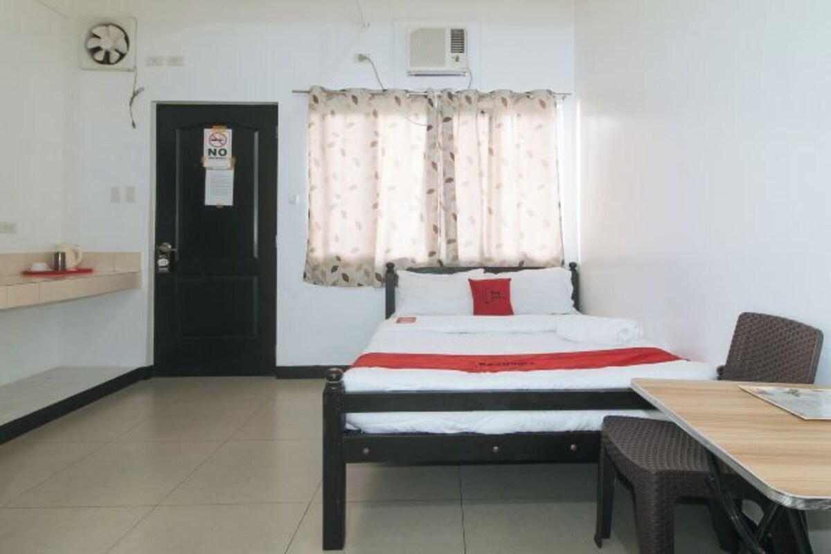 A small, clean room with a bed, a table, and a chair. There is a door with a 'No Smoking' sign, an air conditioning unit, and a fan on the wall.