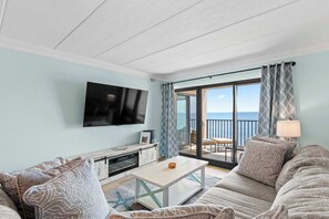 A living room with a TV, sofa, coffee table, and a view of the ocean through a balcony. - Oceanfront Oasis | Indoor Pool | Hot Tub | Deck (Ocean City)