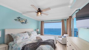 Een slaapkamer met een bed, een plafondventilator, een nachtkastje, een dressoir, een stoel en een raam met uitzicht op de oceaan.
