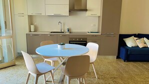 A modern kitchen with white cabinets, a sink, and a stove. There is a dining area with a round table and four chairs. A blue sofa and a small rug are in the living area. - La Casa Sul Pontile (Marina di Vasto)
