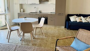 A modern kitchen with white cabinets, a built-in oven, and a sink. There is a dining area with a round table and four chairs. A blue sofa and a wicker chair are in the living area. - La Casa Sul Pontile (Marina di Vasto)