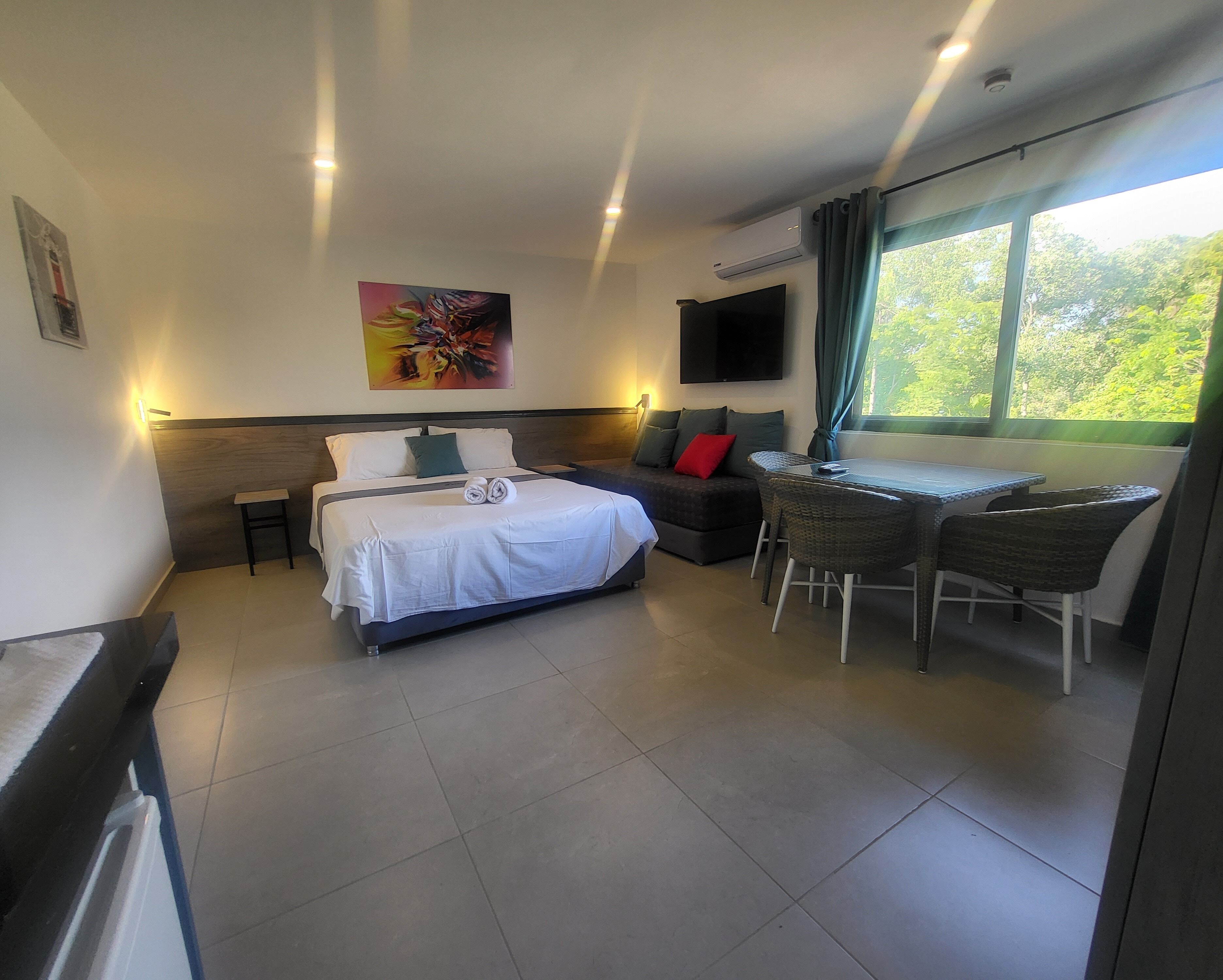 A modern hotel room with a bed, a desk with chairs, a TV, and a painting on the wall.