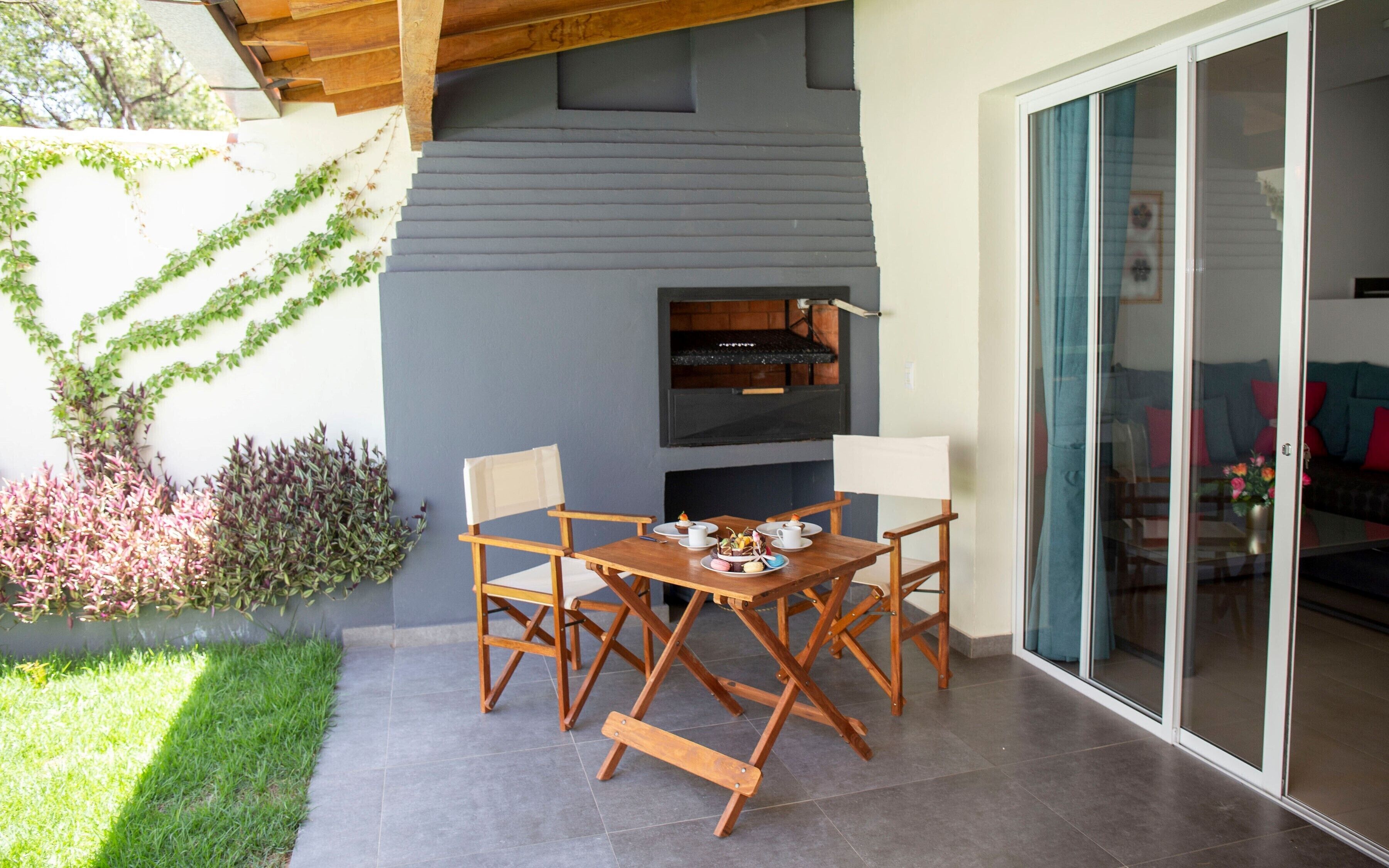 An outdoor dining area with a wooden table and chairs, a built-in grill, and a sliding glass door leading to a living room.
