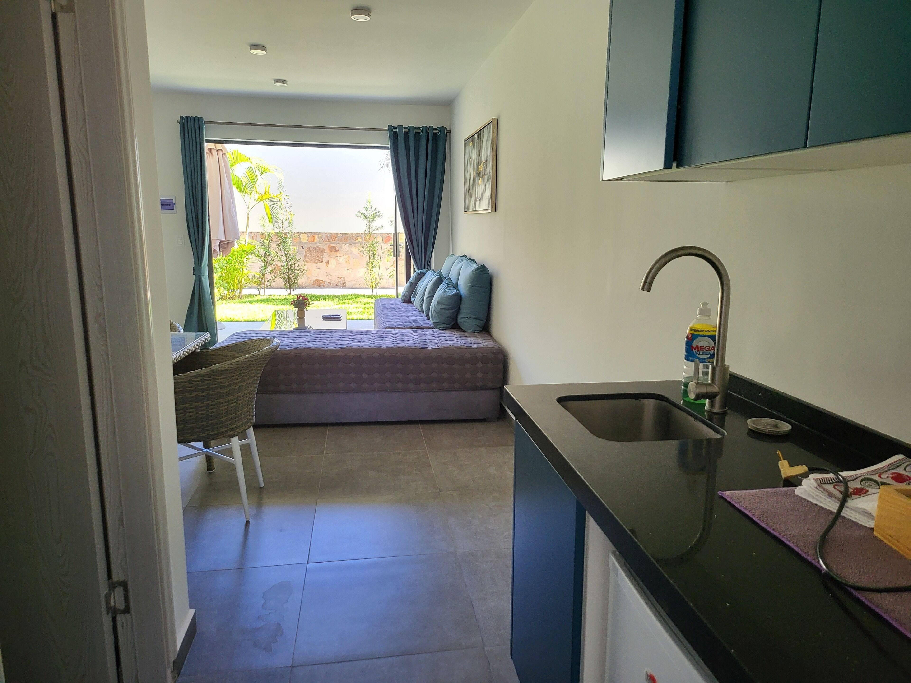 A compact kitchen with a sink, a small dining area with a chair, and a view of a balcony with a bed and curtains.