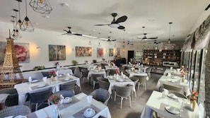 Ein Restaurant mit mehreren Tischen, Korbstühlen, Deckenventilatoren und Wandbildern.