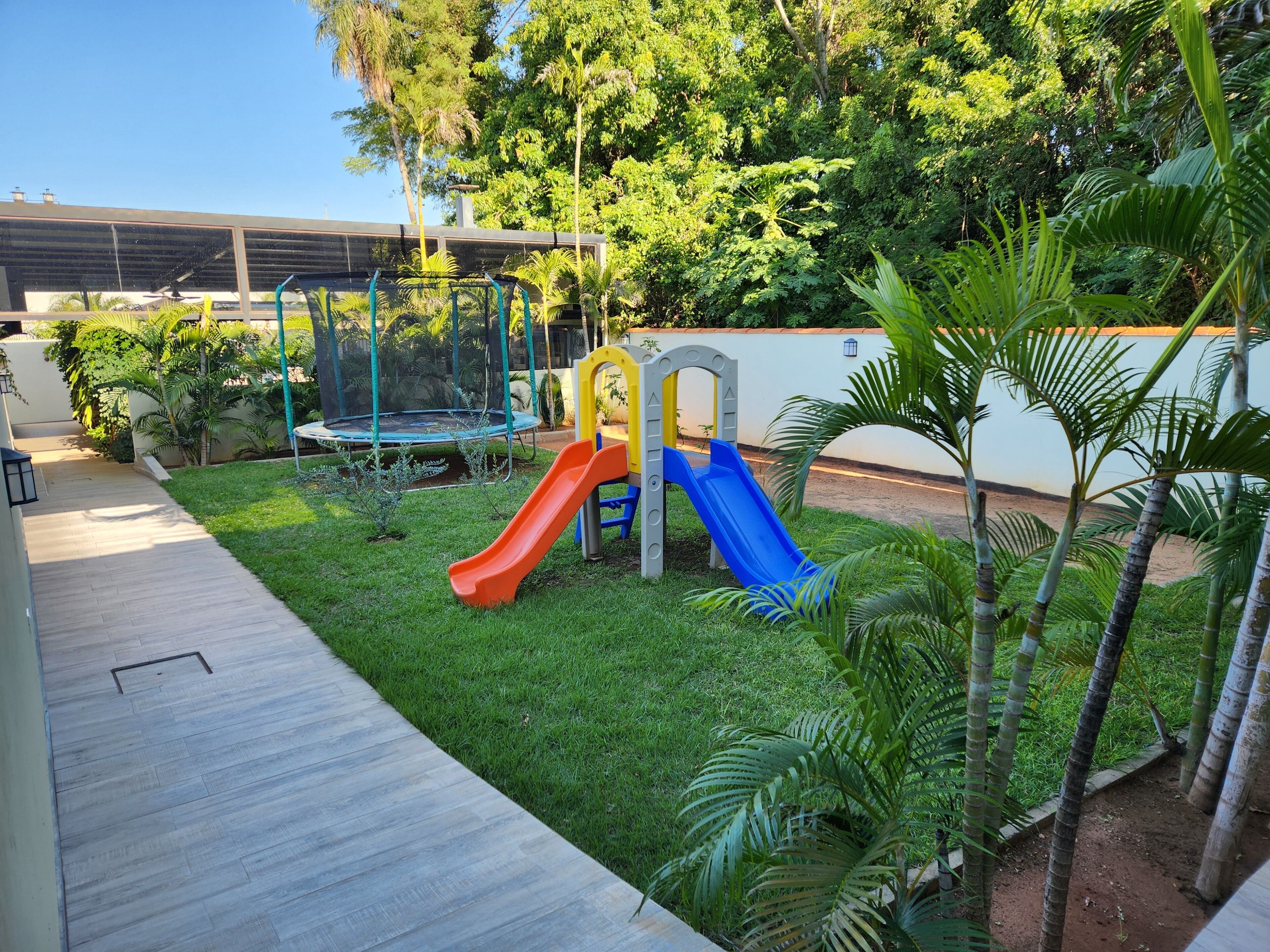 A playground with slides and a trampoline, surrounded by greenery and a paved walkway.