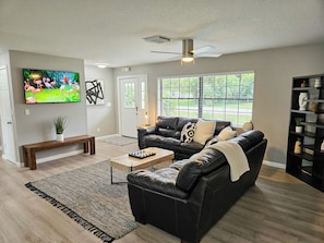 A living room with a black leather sofa, a coffee table, a TV mounted on the wall, a ceiling fan, and a large window with a view of greenery outside.