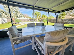A covered patio area with a white table and wicker chairs, overlooking a backyard with a pool and palm trees.