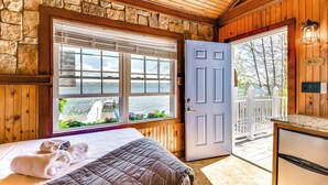 A room with a bed, a window with a view of a lake, a door leading outside, and a stone wall. - Waterfront Romulus Getaway w/ Private Dock! (Romulus)