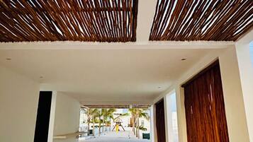 A spacious corridor with a patterned floor, wooden slatted ceiling, and a view of palm trees and a playground in the distance.
