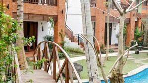 A wooden bridge over a swimming pool, surrounded by a garden with palm trees and a brick building with balconies. - Green Bird Villa (Bali)