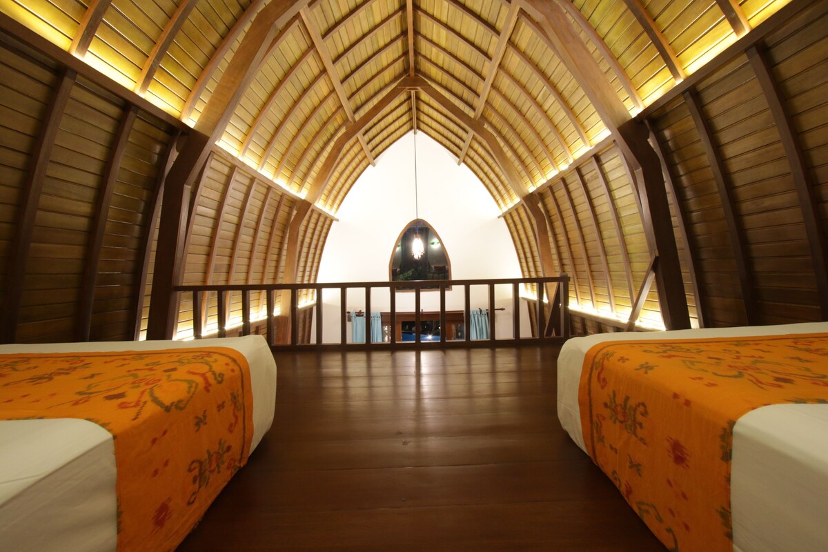 Una habitación espaciosa con techos altos y vigas de madera, dos camas con colchas naranjas y vistas del interior a través de una ventana.
