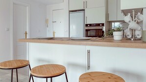 Une cuisine moderne avec des armoires blanches, un réfrigérateur en acier inoxydable, un four et une machine à café. On y trouve deux tabourets en bois et une plante en pot sur le comptoir.