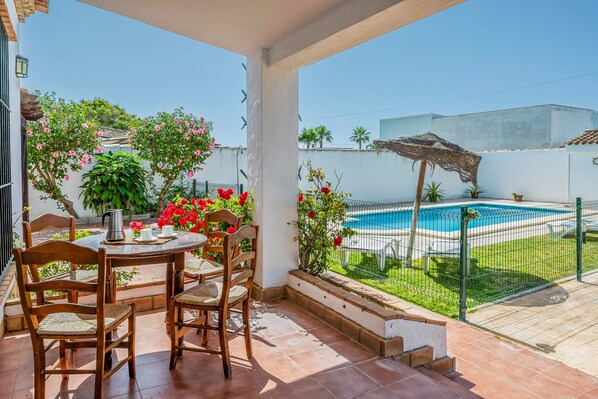 A covered patio with a table and chairs, a pool, and a garden with plants and flowers.