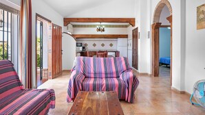 A cozy living area with a sofa, a wooden table, and a kitchen in the background. - Holiday Home 'Chalet Villa Ariza' with Private Pool, Private Garden and Wi-Fi (Cádiz)