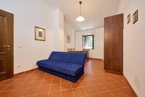 Ein Zimmer mit einem blauen Sofa, einer Holztür, einem Esstisch mit Stühlen und einem Fenster mit Jalousien.