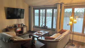 A living room with a sofa, a coffee table, a TV mounted on the wall, and a view of a lake through large windows.