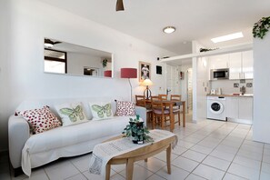 A living room with a sofa, a coffee table, a dining table with chairs, a kitchen area with a microwave, and a washing machine.