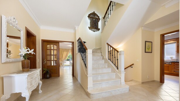 Un hall espacioso con una imponente escalera, un elegante aparador blanco con un jarrón y un espejo grande.