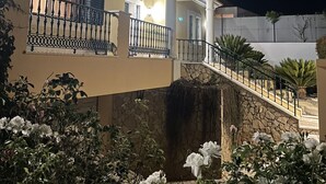 Un edificio de varios pisos con un balcón, una escalera de piedra y un jardín con flores blancas.