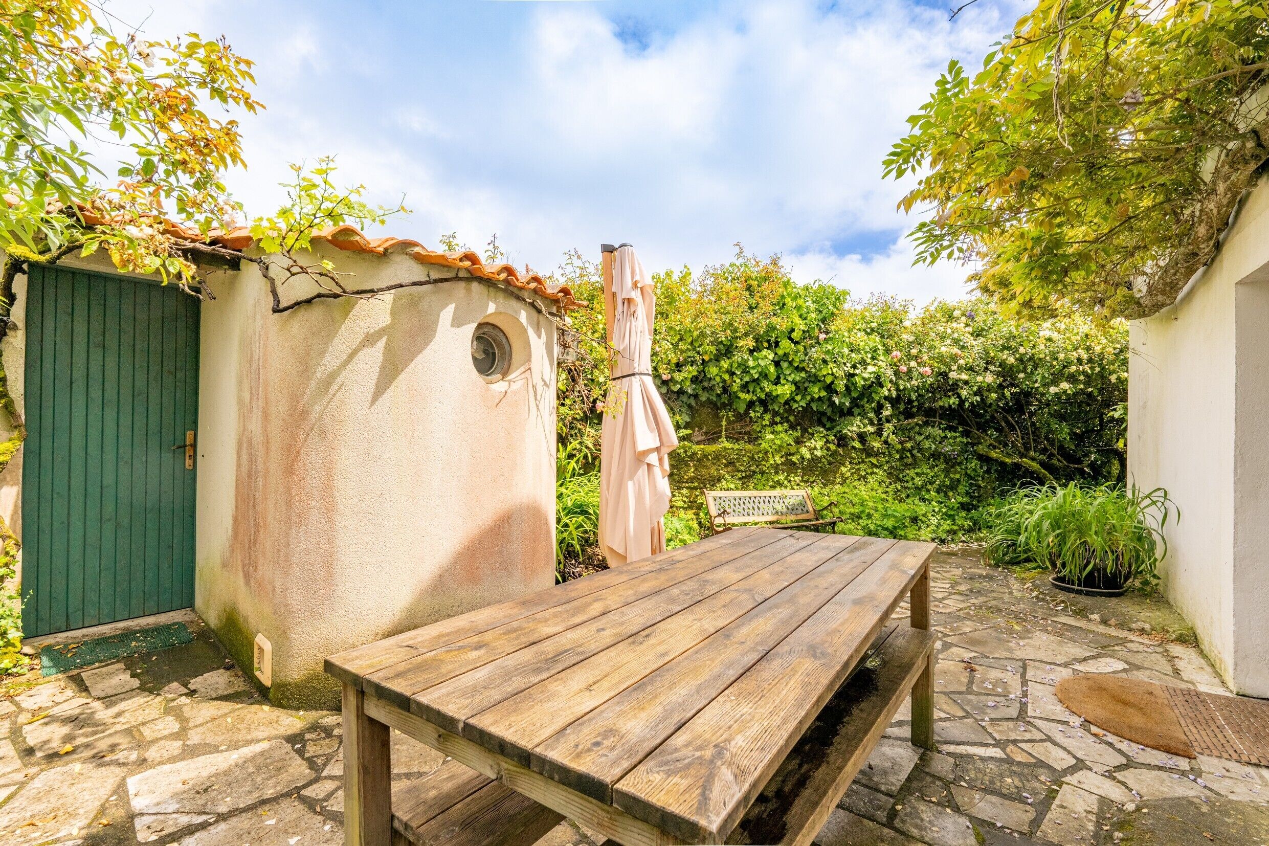 A wooden table in a stone-paved outdoor area with a green door and a small shed.