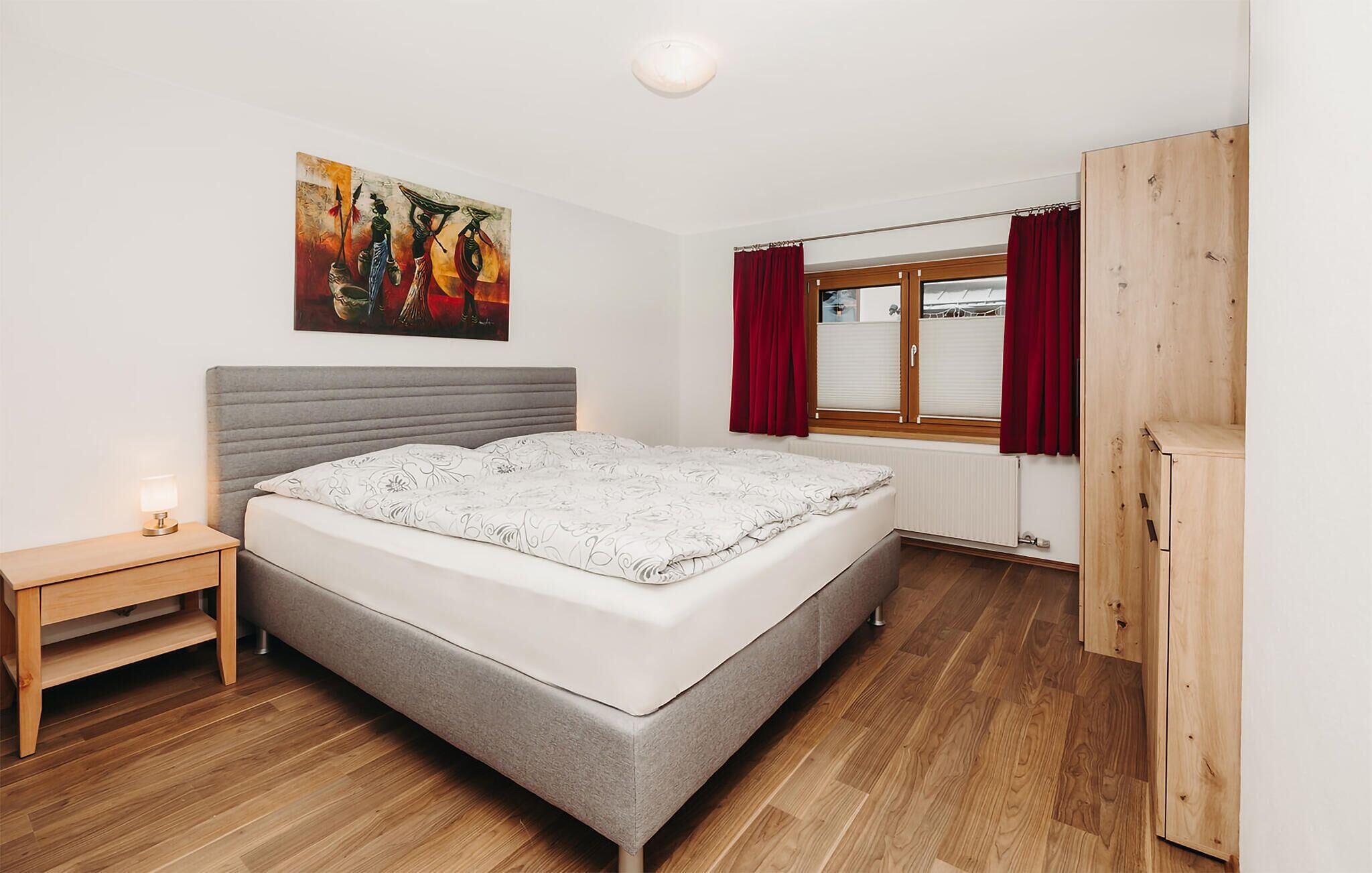 Um quarto com cama, mesa de cabeceira, janela com cortinas e um quadro na parede.