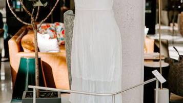 A white dress on a pedestal in a modern interior space with a plant, a sofa, and a table in the background.