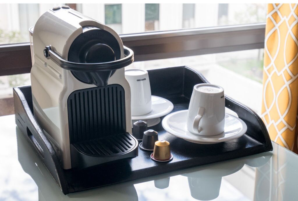 Uma cafeteira sobre uma bandeja com uma chávena e pires, um pequeno recipiente e uma cápsula de café.
