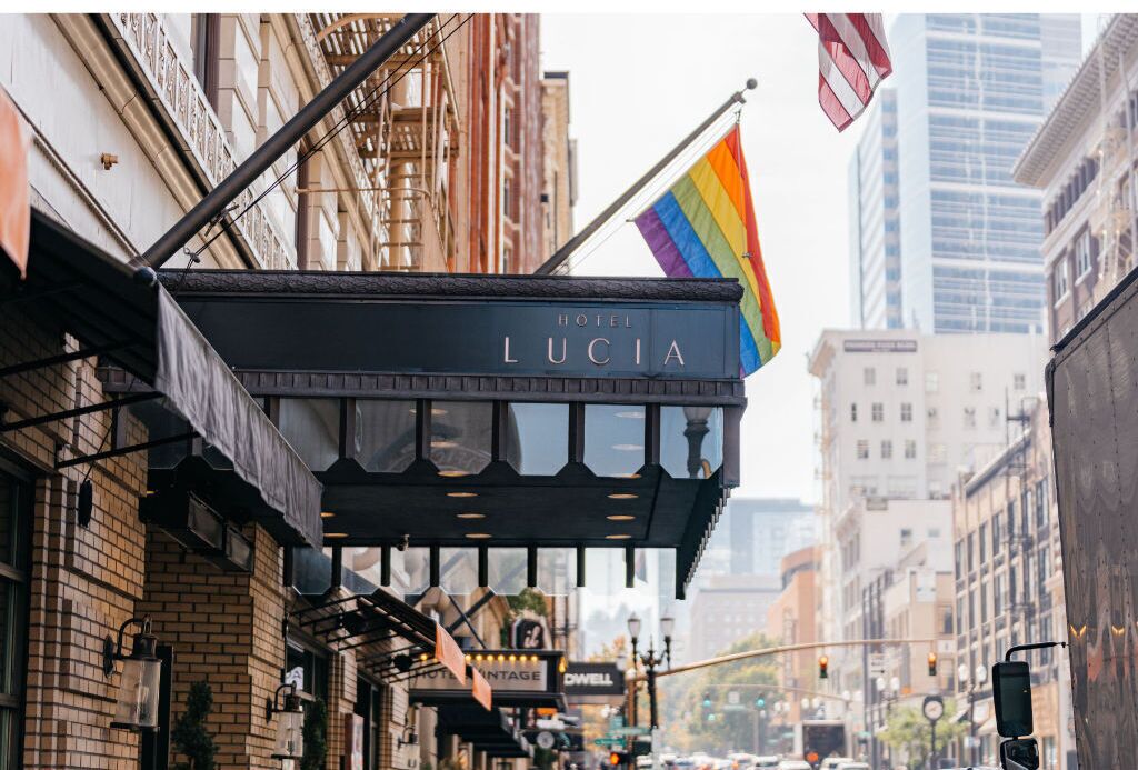 Un hôtel appelé «Hotel Lucia», de style architectural classique, qui se distingue par son auvent imposant et un drapeau arc-en-ciel.