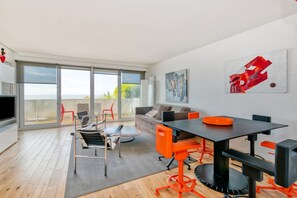 Un salon moderne doté d’une grande porte vitrée donnant sur un balcon, d’un coin repas avec des chaises orange et d’un téléviseur à écran plat.