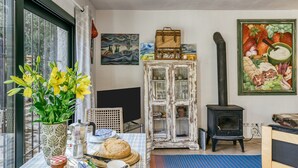 Ein gemütliches Zimmer mit gedecktem Frühstückstisch, einem alten Schrank, einem Kaminofen und Bildern an den Wänden.