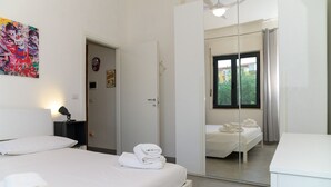 Ein modernes Schlafzimmer mit einem Bett, einer Nachttischlampe, einem Spiegel und Blick auf einen anderen Raum durch eine Glastür.