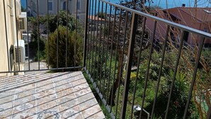 Ein Balkon mit gemusterten Fliesen, einem Metallgeländer und Blick auf eine Küstenlandschaft mit Gebäuden und einem Baum.