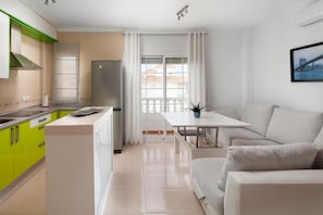 A modern kitchen with green cabinets, a white countertop, and a refrigerator. There is a dining area with a white table and a sofa. - Apartment 'Ático El Morche' with Sea View, Wi-Fi and Air Conditioning (El Morche - Torrox Costa)