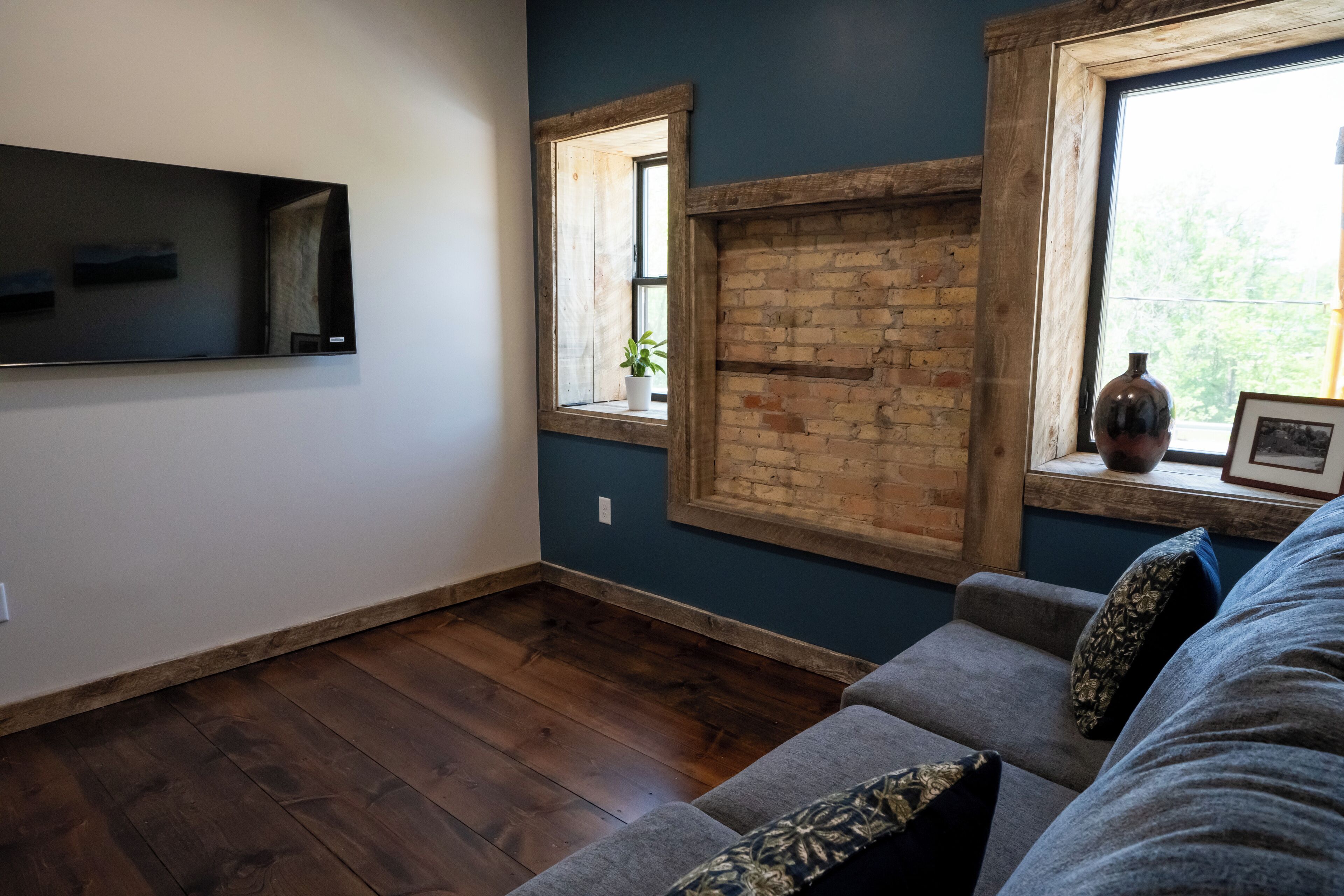 Ein Wohnzimmer mit grauem Sofa, Holzboden, Backsteinwand und einem Fenster mit Pflanze.