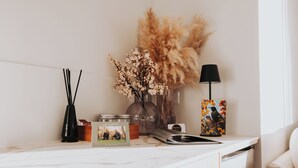 A clean, minimalist kitchen with white cabinetry, a marble countertop, and decorative items including a vase with flowers, a lamp, and a framed picture. - Ahuru House a touch of luxury - Tui Room (Mangawhai)