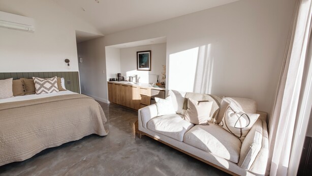 A modern bedroom with a bed, a sofa, and a kitchen area in the background. - Ahuru House a touch of luxury - Tui Room (Mangawhai)