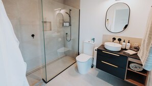 A modern bathroom with a glass-enclosed shower, a white toilet, a sink with a mirror above, and a towel rack. - Ahuru House a touch of luxury - Tui Room (Mangawhai)