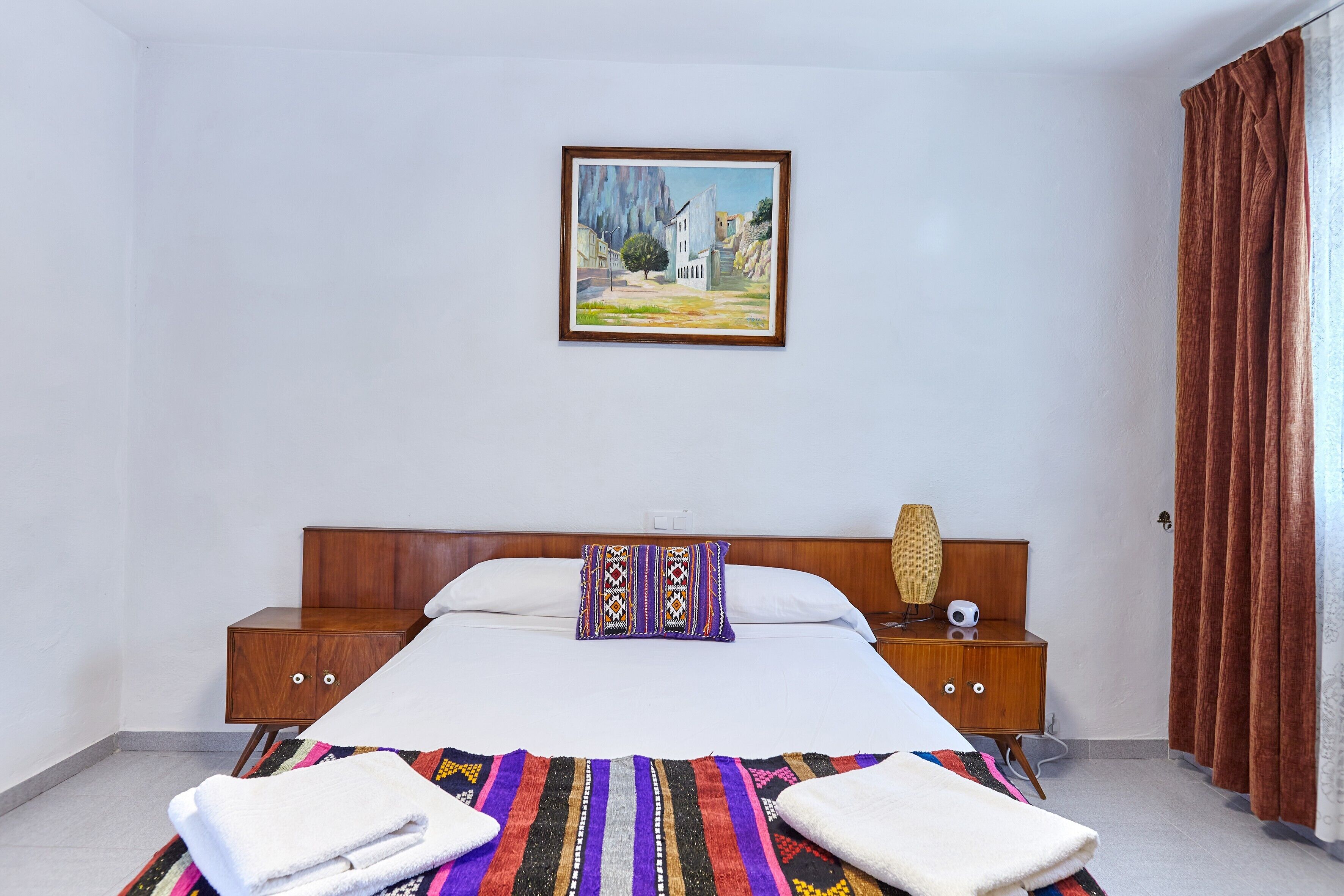 A hotel room with a bed, a nightstand, a lamp, a painting, and a window with curtains.