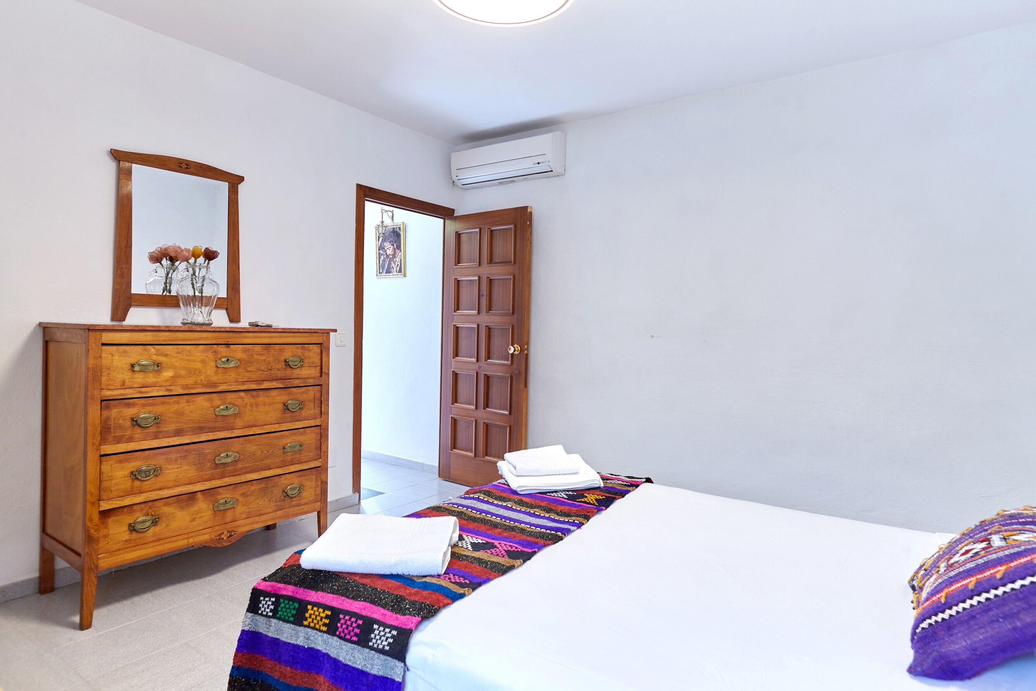 A bedroom with a wooden dresser, a bed with a colorful blanket, and a door leading to another room.