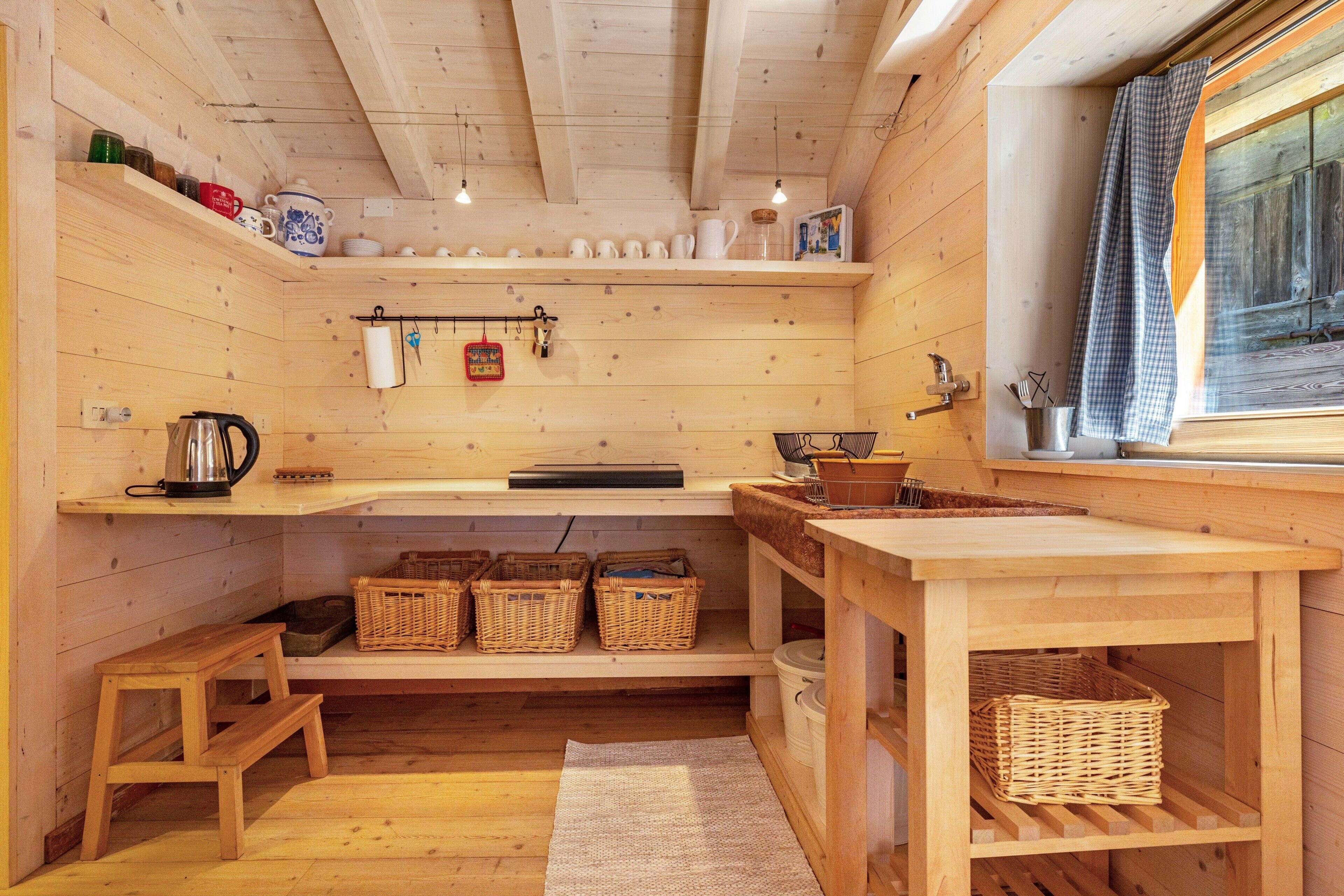 A wooden interior with a kitchen area, shelves with dishes, a kettle, baskets, and a window with a curtain.