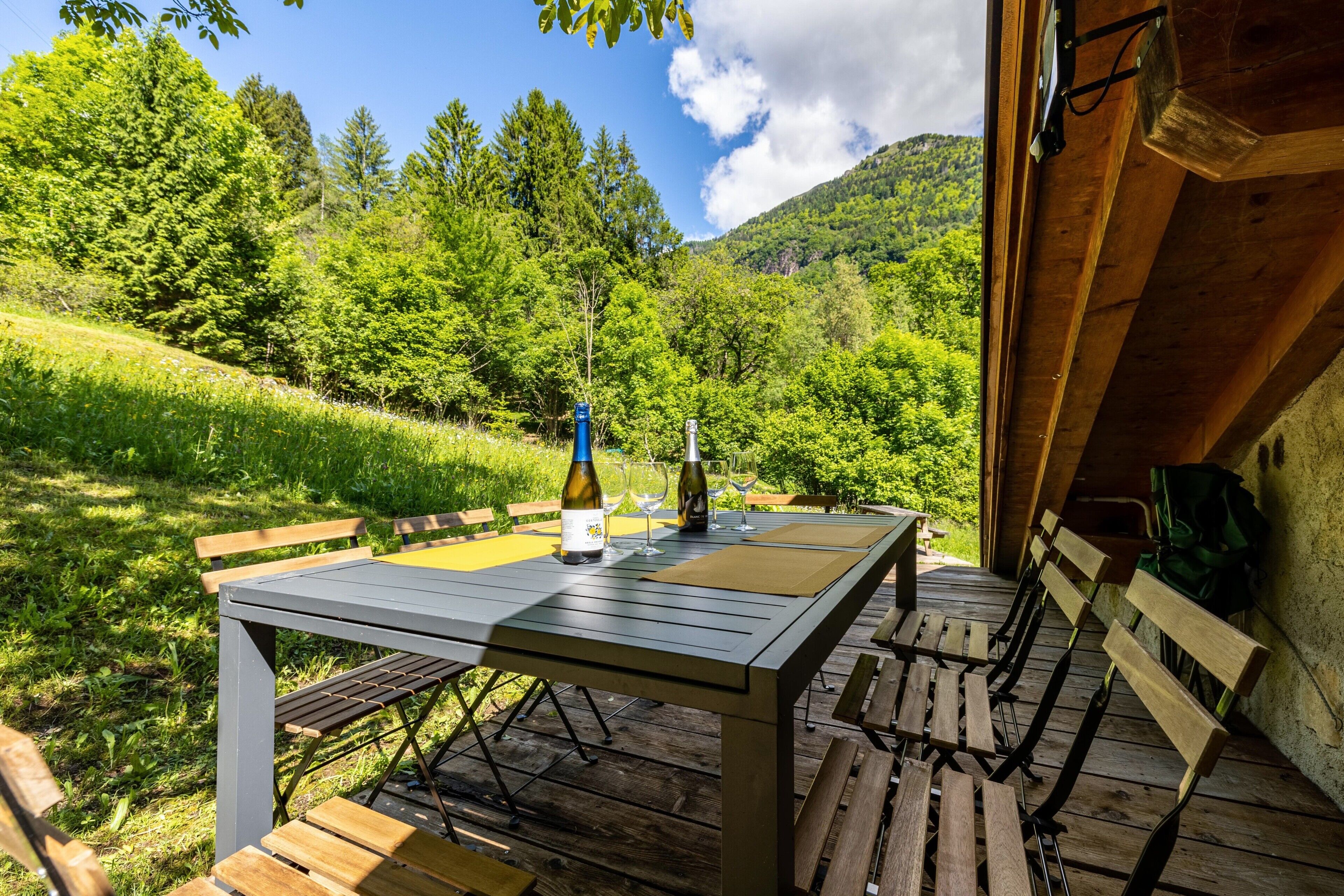 Une table en bois installée à l’extérieur, avec des bancs, une bouteille et des verres, le tout mis en valeur par un paysage verdoyant et luxuriant et une montagne en arrière-plan.